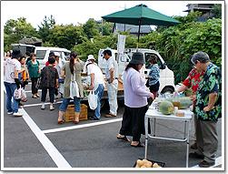 ゆう焼け市で売られている野菜等を手に取ったり見たり買い物をする十数人の人々の写真